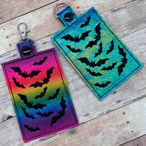Op de afbeelding: Twee sleutelhangers met een regenboog- en blauwe glitterachtergrond en zwarte vleermuis-silhouetten.