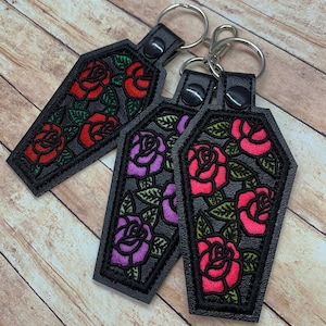 May include: Three black leather keychains shaped like coffins with embroidered roses. One keychain has red roses, one has pink roses, and one has purple roses.