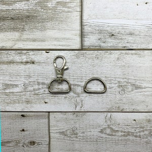Op de afbeelding: Twee zilveren metalen gespen, een met een haak en de andere een D-ring, op een witte houten achtergrond.
