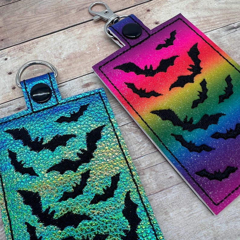 Op de afbeelding: Twee sleutelhangers met zwarte vleermuizen op een regenboog en iriserende achtergrond. De sleutelhangers zijn gemaakt van stof en hebben een metalen ring.