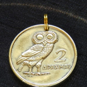 May include: A circular gold-toned metal pendant featuring a relief of an owl on the front. The design includes the number 2 and the Greek text ΔΡΑΧΜΑΙ. A small gold-toned jump ring is attached to the top of the coin-shaped pendant for hanging.