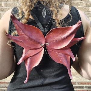 Könnte beinhalten: Burgunderfarbene, blattförmige Flügel, die an einem schwarzen Geschirr befestigt sind. Die Flügel bestehen aus einem strukturierten Material und sind mit silberfarbenen Nieten befestigt. Das Geschirr wird über einem schwarzen Tanktop getragen.