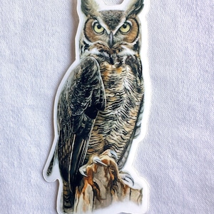 May include: A sticker featuring a detailed illustration of a great horned owl perched on a branch. The owl has brown, gray, and white feathers, with yellow eyes and a sharp beak. The background is a light brown.