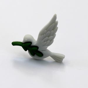 May include: A white dove figurine with outstretched wings, carrying a green olive branch in its beak. The bird is in mid-flight, with detailed feathering. The figurine is a symbol of peace.