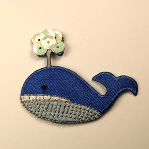 May include: Blue whale applique with a white and green sequin flower on top. The whale has a gray and white textured belly.
