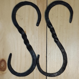 May include: Two handcrafted, dark metal S-hooks. Each hook features a twisted central section and curved ends. The hooks are designed for hanging items and are displayed against a light wood background.