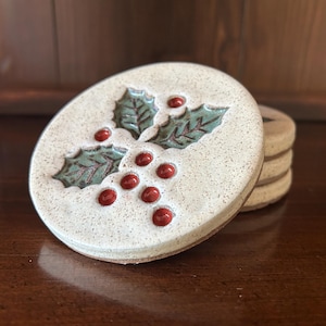 May include: A round ceramic coaster with a speckled off-white glaze featuring a debossed design of green holly leaves and red berries. The coaster has a smooth, glossy finish and is part of a small stack of identical items with a flat circular shape.