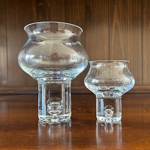 May include: Two clear glass candle holders with a round base and a bulbous top. The larger holder is taller than the smaller holder.