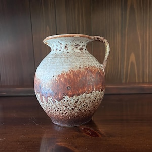 May include: A ceramic pitcher with a handle. The pitcher has a rounded body and a narrow neck. The color is a mix of brown, tan, and white, with a speckled texture. The pitcher is likely used for holding liquids.