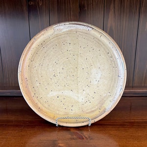 May include: A large, round, ceramic plate with a speckled glaze in shades of beige and brown. The plate has a slightly raised rim and is sitting on a wooden surface.