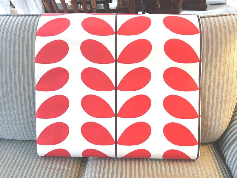 May include: A rectangular pillow with a white background and a repeating pattern of large, red teardrop shapes. The pillow has a black border and is resting on a striped sofa.