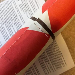 May include: A partially unrolled roll of red and white wallpaper with a brown band. The wallpaper rests on top of a printed instruction sheet. The text "WALLCOVERING HANGING" is visible on the instruction sheet.