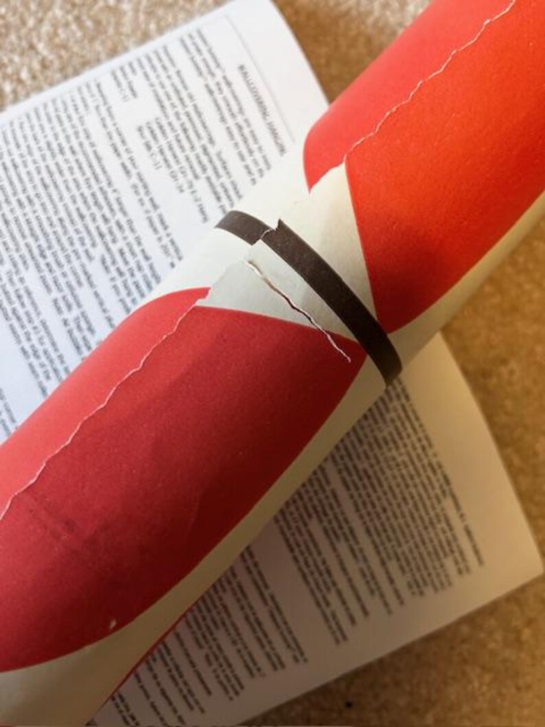 May include: A partially unrolled roll of red and white wallpaper with a brown band. The wallpaper rests on top of a printed instruction sheet. The text "WALLCOVERING HANGING" is visible on the instruction sheet.