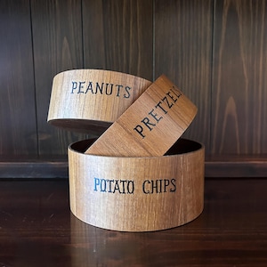 May include: Wooden snack bowls with the words "PEANUTS", "PRETZELS", and "POTATO CHIPS" in black lettering. The bowls are made of light brown wood and are arranged in a decorative display.