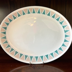 May include: White oval serving platter with a teal and peach fan-shaped border design. The platter is made of ceramic and is displayed on a dark wooden surface. A decorative piece for serving food.