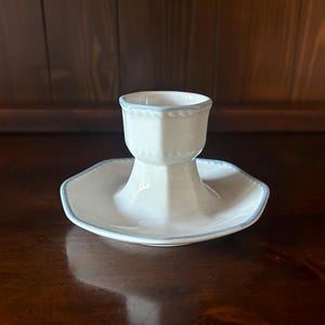 May include: A white ceramic egg cup featuring a hexagonal base and a matching hexagonal rim. The piece is accented with a delicate light blue painted trim along the edges of the base and the top rim, providing a subtle contrast to the white finish.