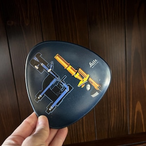 May include: A dark blue, triangular tray with a diagram of a microscope. The diagram features blue and yellow components, with the word "Leitz" in white. The tray is held against a dark wood background.