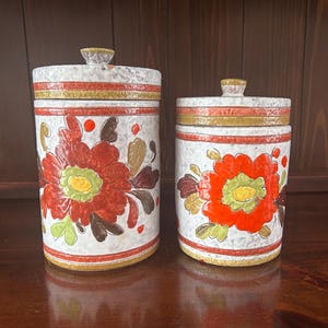 May include: Two ceramic canisters with lids. The canisters are white with a textured finish and decorated with large red and orange floral designs. Gold and brown bands encircle the top and bottom. The lids have small, textured knobs.