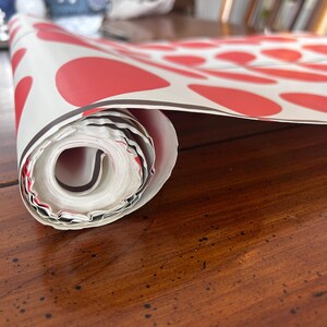 May include: Rolled-up white paper with a repeating pattern of large red circles. The paper has a black border and is resting on a dark wooden surface. The roll is partially unraveled, revealing the inside.
