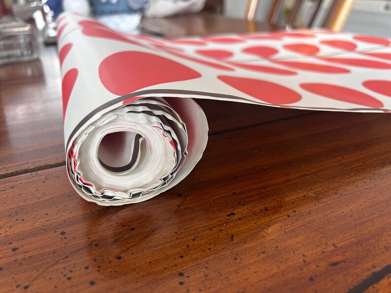 May include: Rolled-up white paper with a repeating pattern of large red circles. The paper has a black border and is resting on a dark wooden surface. The roll is partially unraveled, revealing the inside.