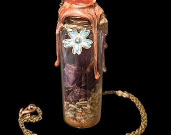 Peut inclure: Une bouteille en verre transparent remplie d'herbes séchées et de pétales violets, scellée avec de la cire brun-rouge et un charm en forme de fleur bleue. Une chaîne dorée est attachée à la bouteille, créant un collier.