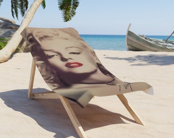 May include: A beach chair with a brown wooden frame and a beige fabric seat featuring a black and white photo of Marilyn Monroe. The photo shows her face with red lipstick and a slight smile.