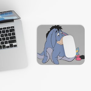 May include: A rectangular mouse mat featuring Eeyore from Winnie the Pooh. The illustration shows Eeyore in a seated position with a sad expression. A white computer mouse rests on the mat. A laptop is partially visible in the background.