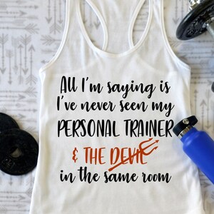 May include: White tank top with the text "All I'm saying is I've never seen my PERSONAL TRAINER & THE DEVIL in the same room". The text is in black with a red devil symbol.