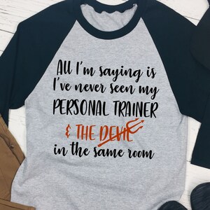 May include: A gray and black baseball tee shirt with the text "All I'm saying is I've never seen my PERSONAL TRAINER & THE DEVIL in the same room".