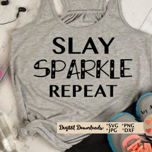 Puede incluir: Un top gris sin mangas con el texto "SLAY SPARKLE REPEAT" en negro. El top está sobre una superficie de madera con una mancuerna rosa, una botella de agua y un par de zapatillas de correr negras y grises.