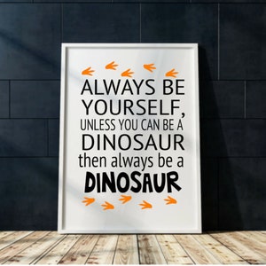 May include: White framed print with black text that reads "Always be yourself, unless you can be a dinosaur, then always be a dinosaur". Orange dinosaur footprints are at the top and bottom of the text.