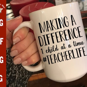 Może przedstawiać: Biały kubek ceramiczny z czarnym napisem "Making a Difference 1 child at a time #TeacherLife".