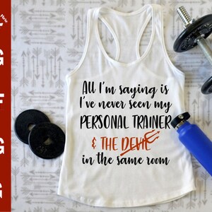 May include: White tank top with the text "All I'm saying is I've never seen my PERSONAL TRAINER & THE DEVIL in the same room". The text is in black with a red devil symbol.