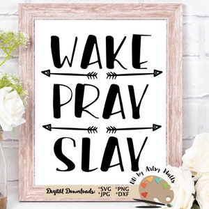 Puede incluir: Una impresión enmarcada con las palabras "WAKE PRAY SLAY" en letras negras en negrita. Hay flechas encima y debajo de las palabras. El marco es de color marrón claro. Flores blancas y vegetación en primer plano.