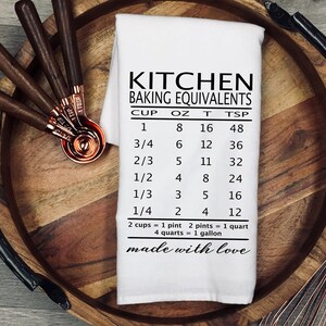 May include: White kitchen tea towel with black text that reads "Kitchen Baking Equivalents" and a chart showing cup, ounce, tablespoon, and teaspoon conversions. The text at the bottom reads "made with love".