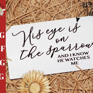 May include: A white card with black calligraphy that reads "His eye is on the sparrow and I know he watches me". The card is sitting on a bed of brown straw.