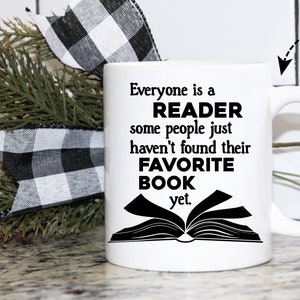 May include: White ceramic mug with a black design that reads "Everyone is a READER some people just haven't found their FAVORITE BOOK yet."  The mug is sitting on a table with a black and white checkered ribbon and a green plant.
