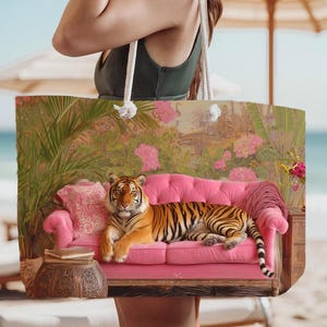 May include: A large tote bag featuring a printed graphic of a tiger resting on a bright pink tufted sofa. The scene includes a patterned pillow, a stack of books on a carved wooden side table, and lush tropical foliage with pink flowers.