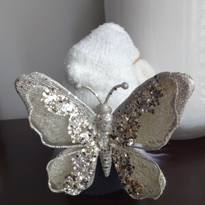Puede incluir: Una mariposa decorativa plateada con detalles de purpurina. La mariposa tiene alas y antenas detalladas. Las alas son de color plata claro con adornos de purpurina y lentejuelas. La mariposa est&aacute; sobre un fondo blanco.