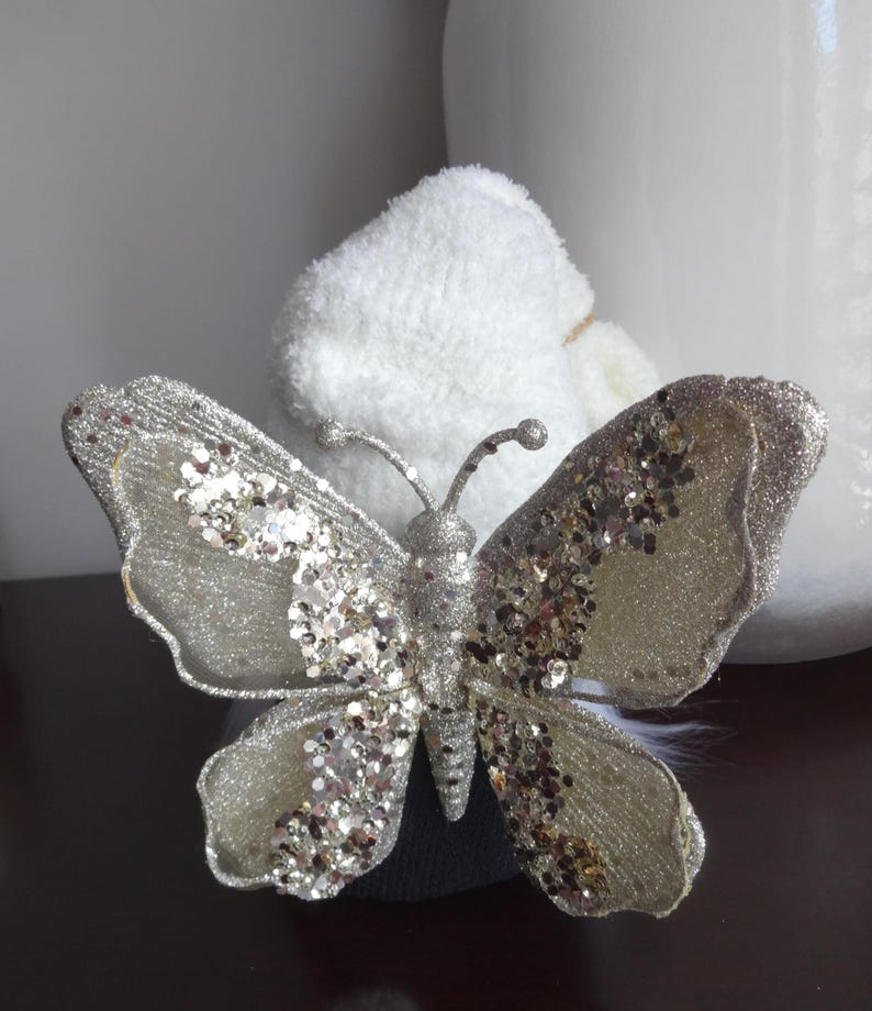 Puede incluir: Una mariposa decorativa plateada con detalles de purpurina. La mariposa tiene alas y antenas detalladas. Las alas son de color plata claro con adornos de purpurina y lentejuelas. La mariposa est&aacute; sobre un fondo blanco.