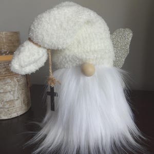 Puede incluir: Figura de gnomo blanco con barba esponjosa, sombrero texturizado y nariz de madera. Tiene un ala peque&ntilde;a de purpurina plateada y un colgante de cruz negra colgando de una cuerda. El gnomo es un art&iacute;culo decorativo.