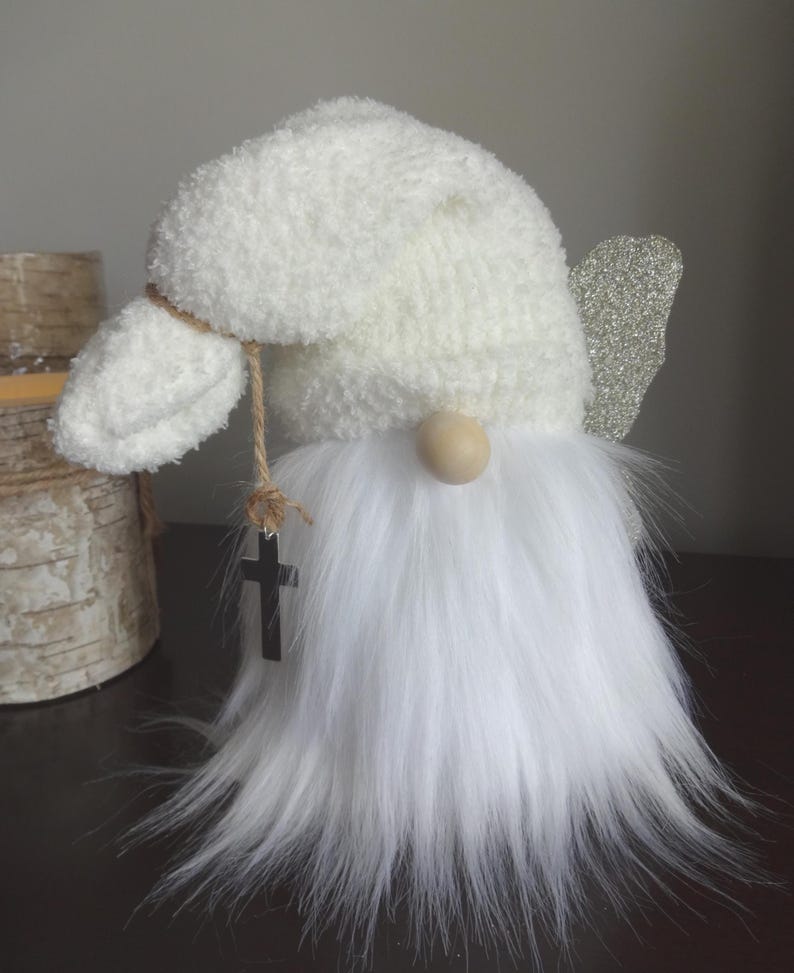 Puede incluir: Figura de gnomo blanco con barba esponjosa, sombrero texturizado y nariz de madera. Tiene un ala peque&ntilde;a de purpurina plateada y un colgante de cruz negra colgando de una cuerda. El gnomo es un art&iacute;culo decorativo.