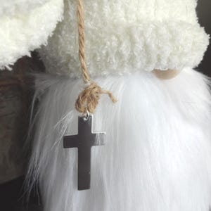 Puede incluir: Un gnomo de peluche blanco con una larga barba blanca y un sombrero a juego. Una cruz plateada cuelga de un collar de cuerda. El sombrero y la barba del gnomo tienen una textura esponjosa.