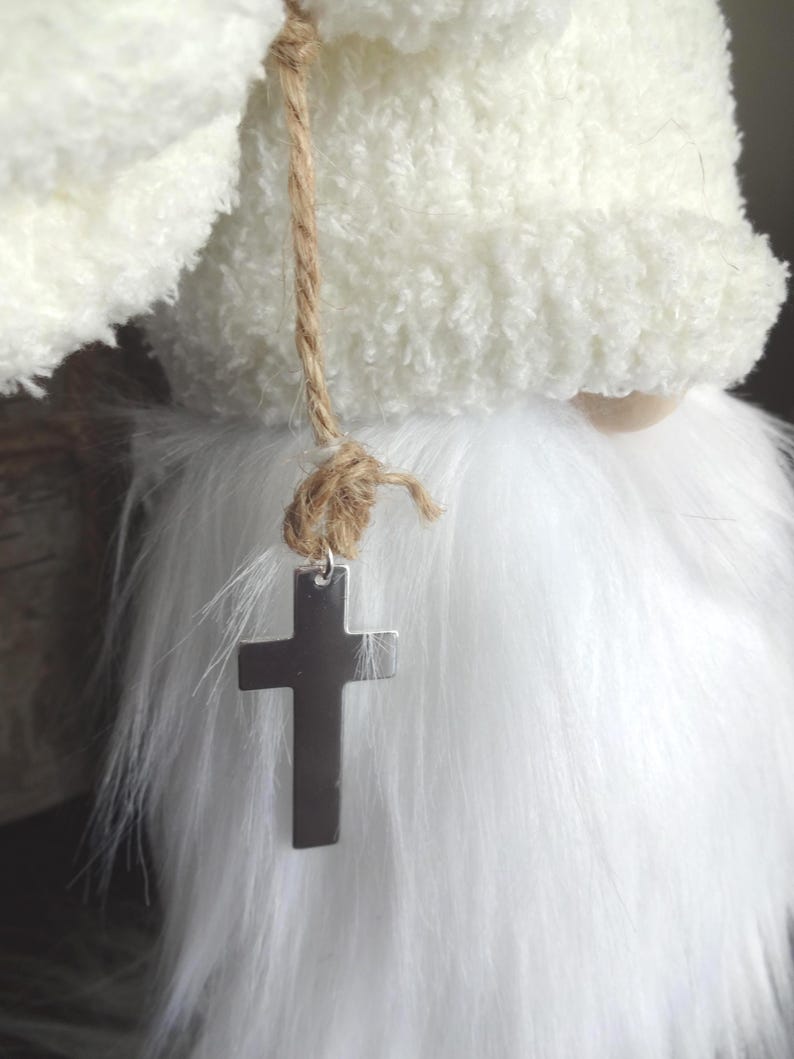 Puede incluir: Un gnomo de peluche blanco con una larga barba blanca y un sombrero a juego. Una cruz plateada cuelga de un collar de cuerda. El sombrero y la barba del gnomo tienen una textura esponjosa.