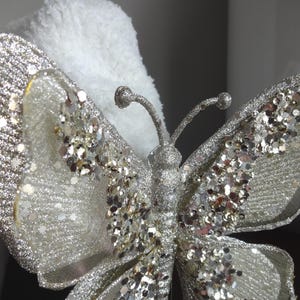 Puede incluir: Primer plano de una mariposa decorativa con alas plateadas brillantes y cuerpo plateado. La mariposa tiene una delicada textura similar a una malla. El fondo es blanco y esponjoso.