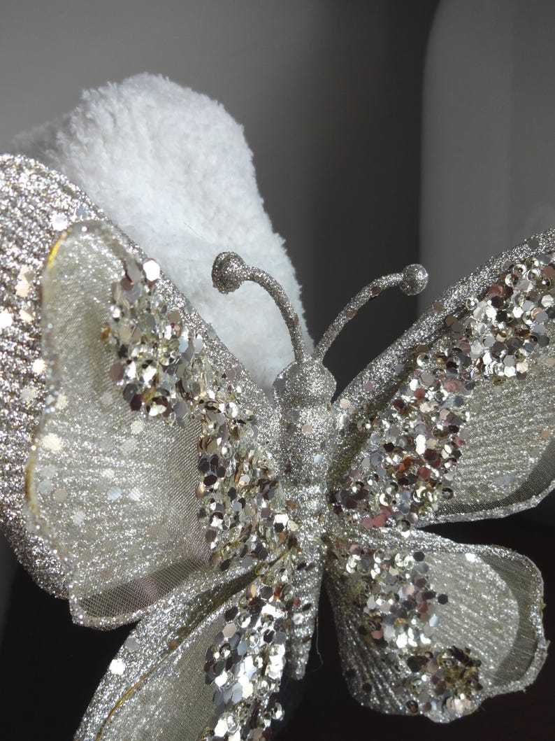 Puede incluir: Primer plano de una mariposa decorativa con alas plateadas brillantes y cuerpo plateado. La mariposa tiene una delicada textura similar a una malla. El fondo es blanco y esponjoso.