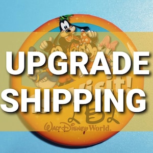 May include: A yellow and orange round button with a rainbow arc and the text "Visit!" in a white circle. The button features cartoon characters Mickey Mouse, Minnie Mouse, Pluto, and Goofy. The text "Upgrade Shipping" is superimposed on the button. The text "Walt Disney World." is at the bottom of the button.