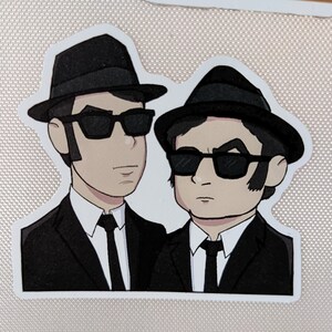 May include: A sticker featuring a cartoon illustration of two men in black suits, hats, and sunglasses. The figures are side-by-side, with a white border around the image. The sticker is likely a pop culture reference.