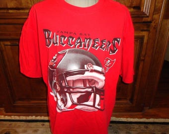 Puede incluir: Camiseta roja con un gráfico de un casco de los Tampa Bay Buccaneers y el nombre del equipo "Tampa Bay Buccaneers" en una fuente estilizada.