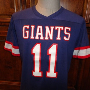 May include: A blue football jersey with white and red trim. The jersey has the word "GIANTS" in white letters with red outlines and the number "11" in white letters with red outlines.
